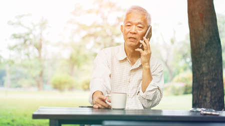 Asian senior man talking on phone white drinking coffee in parkの写真素材