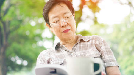Asian senior woman reading book drinking coffee in sunny parkの写真素材
