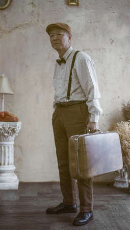 Retro vintage fashion Asian senior man with flat cap and suspender carry luggage for travel lifestyleの写真素材