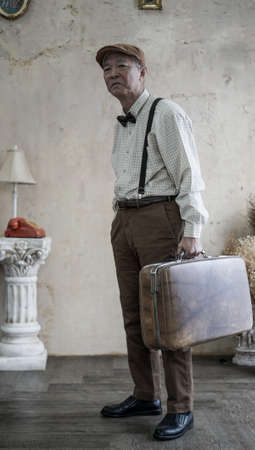 Retro vintage fashion Asian senior man with flat cap and suspender carry luggage for travel lifestyleの写真素材