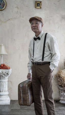 Retro vintage fashion Asian senior man with flat cap and suspender carry luggage for travel lifestyleの写真素材