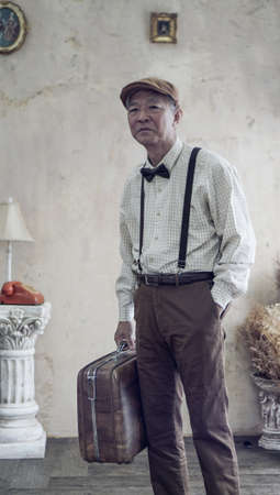 Retro vintage fashion Asian senior man with flat cap and suspender carry luggage for travel lifestyleの写真素材