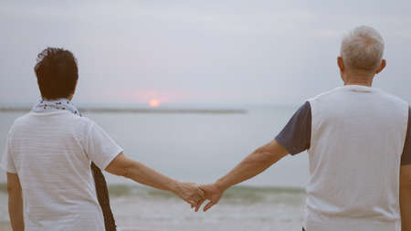 Asian elder senior couple holding hands looking sunset sea ocean together happy retirement lifeの写真素材