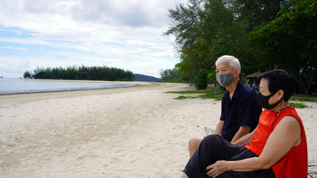 Asian senior couple with mask relax sitting on beach log white sand landscape beautiful seaの写真素材