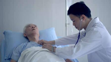 Asian doctor checking heart and pulse condition on senior in hospital bed with stehoscopeの写真素材