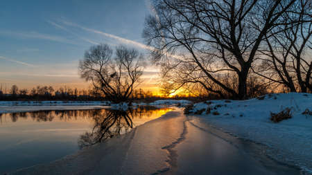 Scenic evening winter landscape. beautiful glow Dramatic sunset over the river to the coast suburb under snow and ice edge. February thawの写真素材