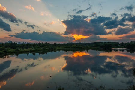 A beautiful summer evening. The dramatic sunset reflects in the smooth water of the river or lake on the urban outskirtsの写真素材