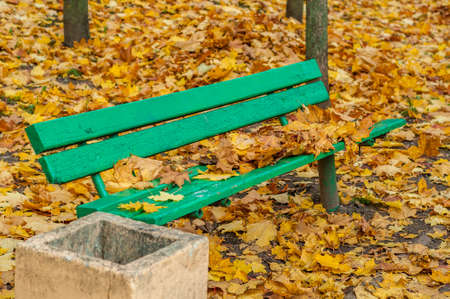 old wooden bench in an autumn park is covered with leaves against a background of yellow fallen maple leavesの写真素材