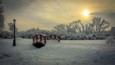romantic winter landscape. A picturesque evening city park covered with snow in cloudy weather. a footpath with a small wooden bridge and a street lamp against the sky with an orange-yellow tingeの写真素材