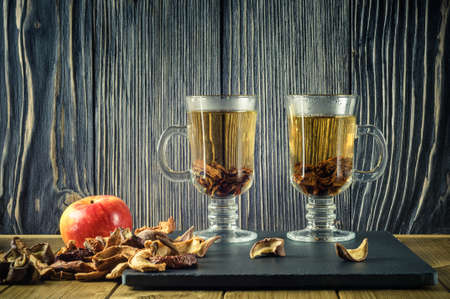 two clear glass cups of hot tea with dried fruit and fresh apple on a stone plate and a wooden backgroundの写真素材
