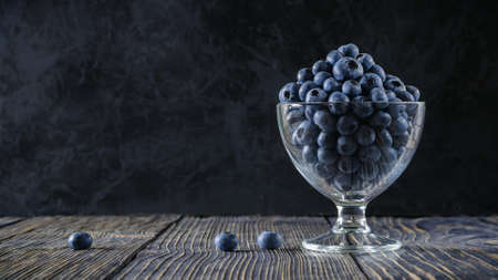 Ripe garden large blueberries in a glass bowl on the right on an old textured wooden table against a background of black concrete plaster with a muffled mystical light. side view. dark photoの写真素材