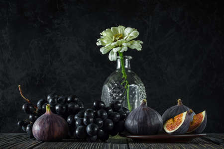 ripe whole and sliced figs with a bunch of black grapes and one flower in an empty decanter on old wooden boards against the background of a black textured concrete wall. elegant classic still life in the style of a dark photo. side viewの写真素材
