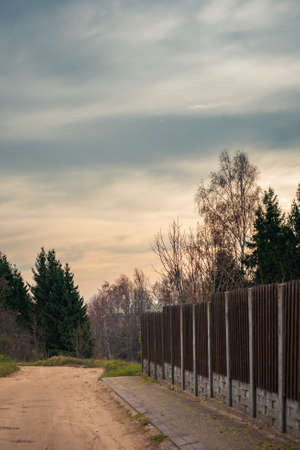 a deserted rural dirt road in a village with a roadside paved with concrete tiles along a modern fence and trees under a beautiful cloudy skyの写真素材