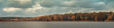 late fall variegated colors. wide panoramic view across the water surface to the opposite river bank with coastal forest under a cloudy sky on October overcast day. colorful autumn landscapeの写真素材