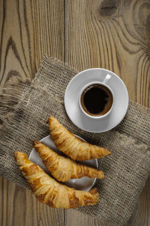 three fresh homemade croissants on a saucer and a cup of hot black coffee on a rough burlap napkin on a wooden textured background. top view. stylish art picture with copy spaceの写真素材