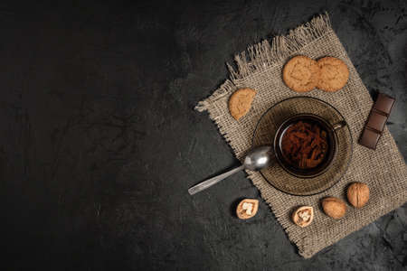 tea in a transparent cup on a saucer with a spoon, with cookies, walnuts and chocolate on a rough burlap napkin and a black plastered concrete background. top view with copy spaceの写真素材