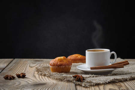 hot coffee in a white cup with light steam, muffins, cinnamon sticks and star anise on a rough burlap napkin on a wooden table against a black concrete wall. artistic mockup with copy spaceの写真素材