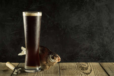 a full misted glass of dark cold foamy beer with dried river fish on rough boards against a black...