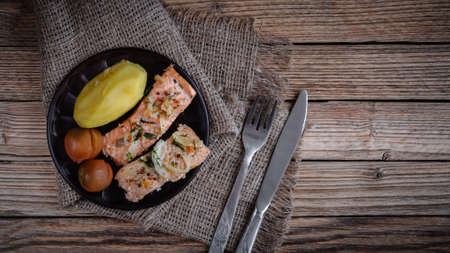 slices of baked salmon with boiled potatoes and pickled tomatoes on a clay plate on a burlap napkin with cutlery on old textured wooden boards. top view. artistic rustic layout with copy spaceの写真素材