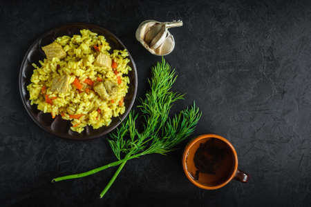 freshly prepared pilaf with meat on a clay plate with a cup of tea, a bunch of fresh dill and a head of garlic on a black concrete textured background. top view. Elegant creative artistic mockup with copy spaceの写真素材