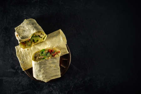 whole and half-cut shawarma kebab stuffed with chicken, vegetables and sauce on a round clay plate on a textured dark concrete plaster background. top view.creative artistic mockup with copy spaceの写真素材