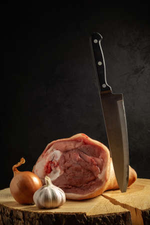 raw pork knuckle on a cracked wooden plate with onions, garlic and a vertically thrust chopping knife on a black concrete background in semidarkness. moody picture with copy space for creative mockupの写真素材