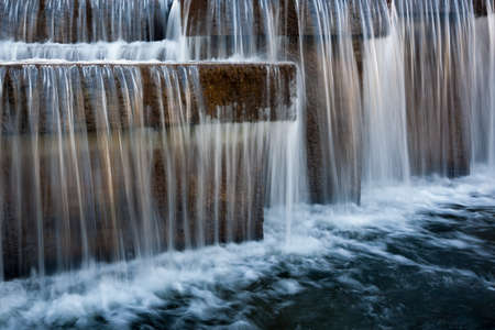 beautiful cascading artificial waterfall from stone and concrete blocks on the city water river channel with blurry streams and a bubbling foamy surface of the water. close-up side viewの写真素材