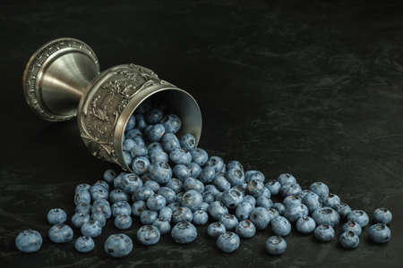 ripe fresh blueberries spilled out of a lying antique silver glass onto the black concrete surface. side view. atristic minimalist moody still life with copy spaceの写真素材