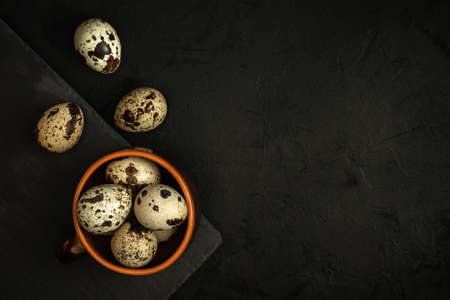 raw quail eggs in a clay cup on the edge of a black stone cutting board on a textured dark concrete surface. moody artistic mockup with copy space. top viewの写真素材