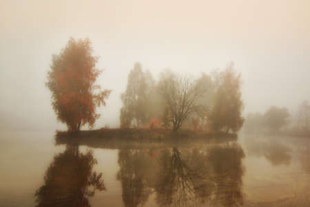 dull autumn landscape. a small island with trees and dry grass in dense fog in the middle of a lake with reflection on the smooth surface of the water. sad colors of late autumnの写真素材