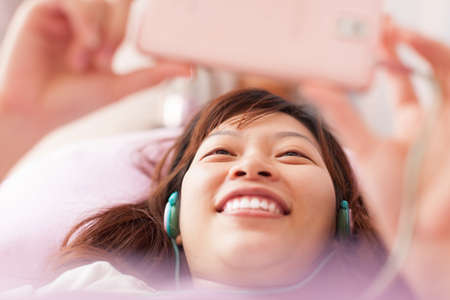 Asian young girl using smart phone wear headphones lying on bedの写真素材