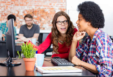 Two colleagues woman talking discussing sitting office desk computer business people working casual...