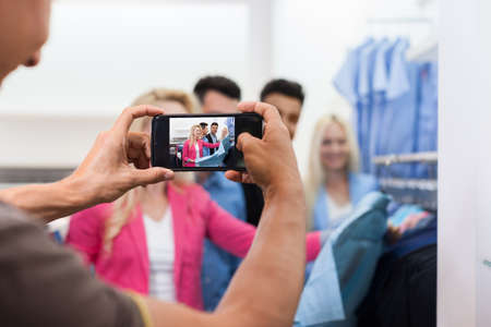 Man Taking Photo Of Young People Shopping, Happy Smiling Friends Two Couple Customers In Fashion Shop Choosing Clothes Shirtsの写真素材