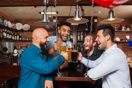 Man Group In Bar Clink Glasses Toasting, Drinking Beer Hold Mugs, Mix Race Cheerful Friends Wear Shirts Meeting Pub Communicate Talkingの写真素材