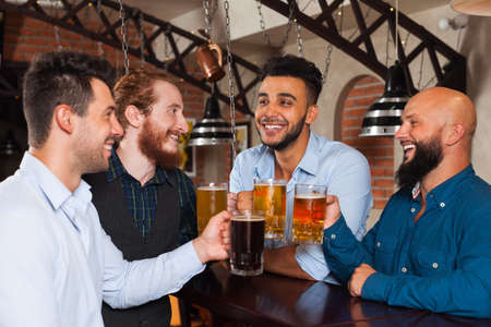 Man Group In Bar Clink Glasses Toasting, Drinking Beer Hold Mugs, Mix Race Cheerful Friends Wear Shirts Meeting Pub Communicate Talkingの写真素材