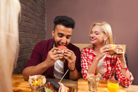 Young Man And Woman Eating Fast Food Burgers Sitting At Wooden Table In Cafe, Friends Meeting Communicationの写真素材