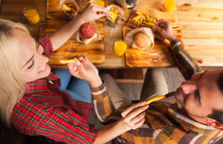 Young Man And Woman Eating Fast Food Potato Sitting At Wooden Table In Cafe Top Angle View, Friends Meeting Communicationの写真素材