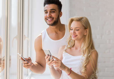 Young Beautiful Couple Stand Near Big Window, Using Cell Smart Phone Happy Smile Hispanic Man Woman...