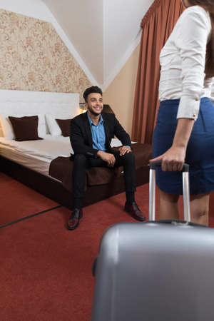 Latin Businessman Sitting At Bed In Room Businesswoman With Luggage Suitcase Business Man And Woman Arriving To Hotelの写真素材