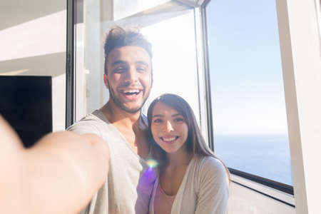 Young Couple Taking Selfie Photo Embracing Asian Woman And Hispanic Man Happy Smiling Modern Apartment With Big Windows Interiorの写真素材