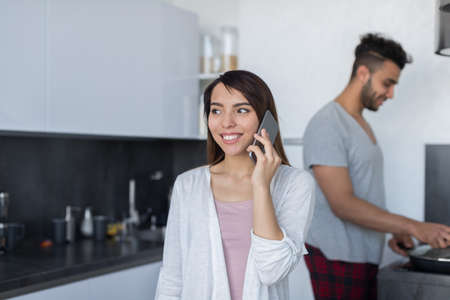Young Couple In Kitchen, Asian Woman Talking Phone Call, Hispanic Man Cooking Breakfast Modern Apartment Interiorの写真素材