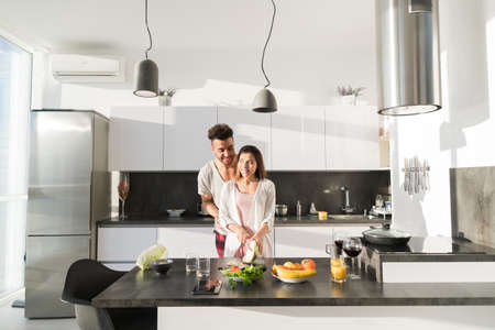 Young Couple Embrace In Kitchen, Hispanic Man And Asian Woman Hug Modern Apartment Interior