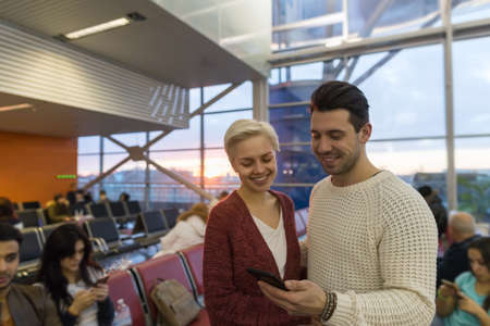 Young Couple In Airport Lounge Waiting Departure Happy Smile Man And Woman Use Cell Smart Phone, Flight Delayの写真素材