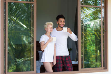 Young Couple Drink Coffee Or Tea, Man And Woman Awake Enjoy Tropical Forest View From Bedroom Hotel...