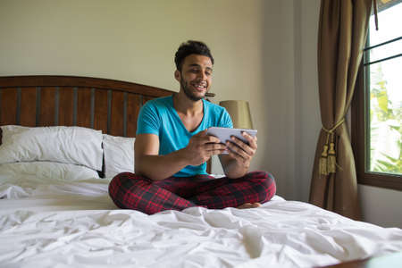 Young Man Sit On Bed, Happy Smile Hispanic Guy Bedroom Using Cell Smart Phone Hotel Room Interiorの写真素材