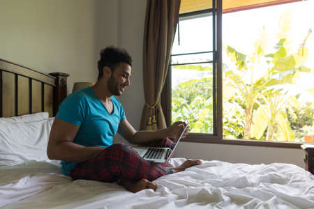 Young Man Sit On Bed, Happy Smile Hispanic Guy Bedroom Using Laptop Computer Hotel Room Interiorの写真素材