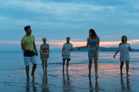 People Talking On Beach At Sunset, Young Tourist Group Walking On Sea In Evening Communication While Summer Vacationの写真素材
