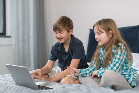 Small Boy And Girl Use Laptop Computer Sitting On Bed In Bedroom, Brother And Sister Surfing Internet Togetherの写真素材