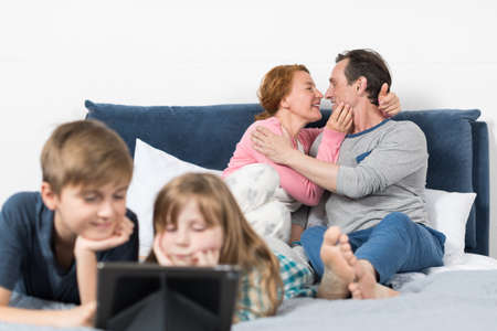 Children Watching Movies On Tablet Computer Lying On Bed Over Embracing Parents Background In Bedroomの写真素材