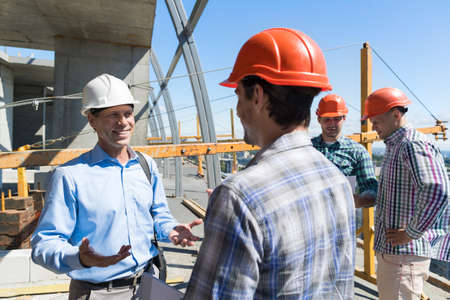 Builders Meeting On Construction Site Architect Talking With Contractor Over Group Of Apprentice...